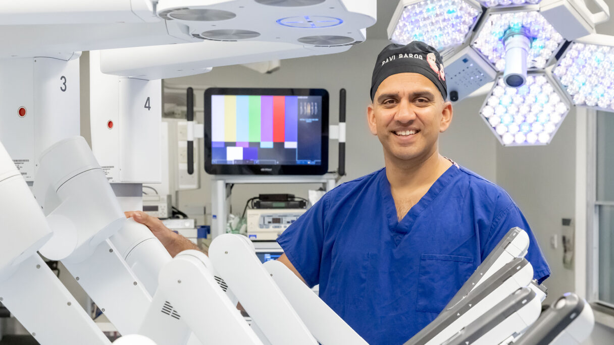 A man in medical scrubs and cap in a hospital clinical ward standing behind a surgical robot with multiple arms.