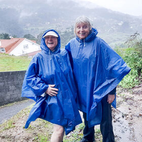 Two women in blue rain ponchos, they are on the Camino trail. There are mountains in the background, it is muddy and raining.