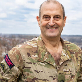 A man wearing army clothing smiling while standing on a rooftop of a tall building in the middle of the day.