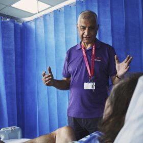 A man in a Royal Free Charity volunteer t-shirt with his arms out wide, speaking to a patient in a hospital ward.