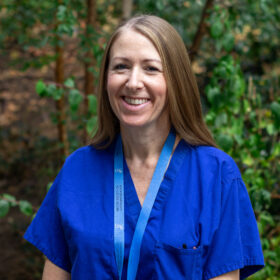 A person with long light brown hair is wearing blue medical scrubs and a blue lanyard standing outdoors in front of leafy green foliage.