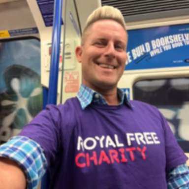 Man stood in a Tube carriage smiling while wearing a purple t-shirt that says Royal Free Charity