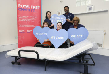 Five people holding large blue hearts - with words on them - standing and kneeling in front of a portable bed. Beside them all is a Royal Free Charity banner.