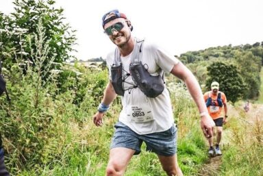 Man in full running gear and sunglasses smiling and running through the field with another runner behind him.