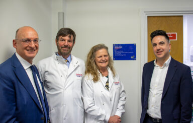 Four people standing in a row, two wearing Royal Free Charity lab coats, by a plaque on the wall that reads: Miranda Filmer neuroendocrine cancer research funding for Spatial Biology Hub.