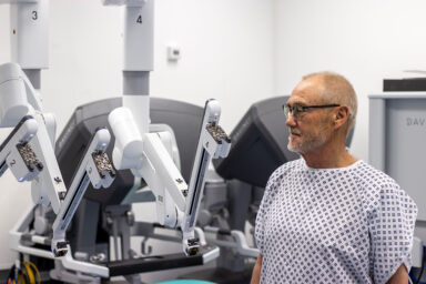 A patient in a hospital gown and glasses standing beside the new surgical robot.