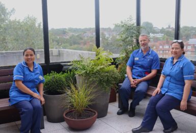 Three members of NHS wearing blue uniforms sat on benches in an outdoor garden.