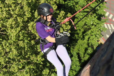 A woman wearing a Royal Free Charity t-shirt, abseiling down the Pears Building. The background is green leaves on a tree.
