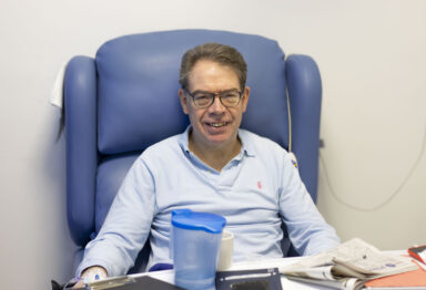 A man sat in a blue hospital chair with a desk in front of him. He is wearing a blue, long-sleeved t-shirt and glasses and is smiling.