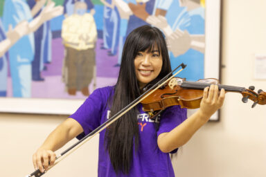 A woman with long dark hair wearing a Royal Free Charity t-shirt is playing the violin and smiling at the camera.