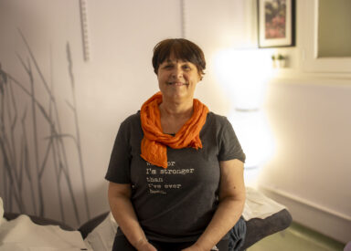 A woman with short brown hair smiles while sitting cross-legged on a bed or cushion in a softly lit room. She is wearing a dark grey t-shirt with text and an orange scarf around her neck.