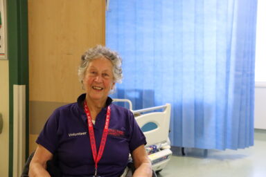 A woman who is wearing a purple Royal Free Charity tshirt while sat in a wheelchair smiling.