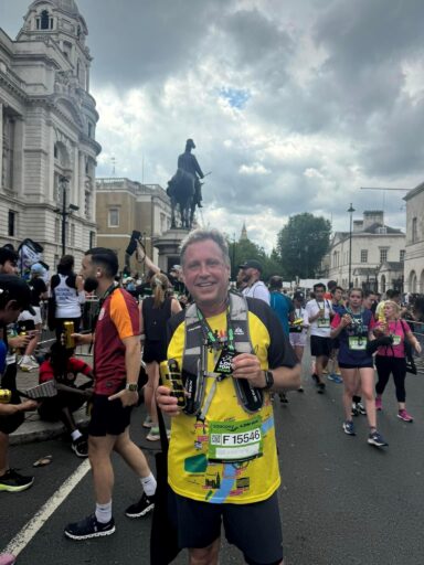 Man standing in the rood in running gear wearing a finishes medal, surrounded by other runners.
