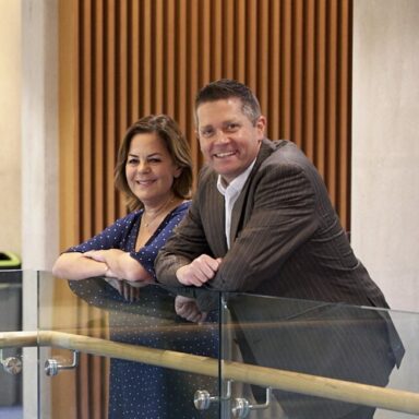 A smartly-dressed woman and man are leaning on a glass railing, smiling at the camera.