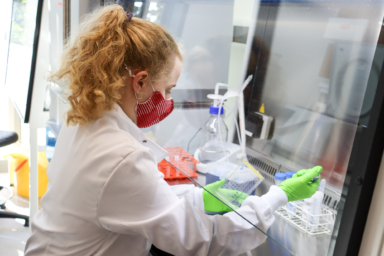 A female reasearcher working at a station within the UCL lab. She is wearing a white lab coat with green latex gloves.