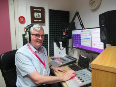 A man sat at a radio desk with a computer and microphone in front of him.