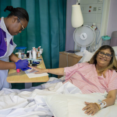 A female beautician, who is wearing a Royal Free Charity branded t-shirt, painting the nails of a woman who is lay in a hospital bed.