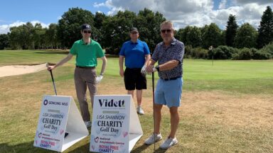 Three men on a golf course holding golf clubs. There are two signs that say 'Lisa Shannon charity golf day'.