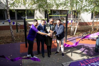 Four people standing in a row holding large scissors and cutting a ribbon in front of some outdoor gym equipment.