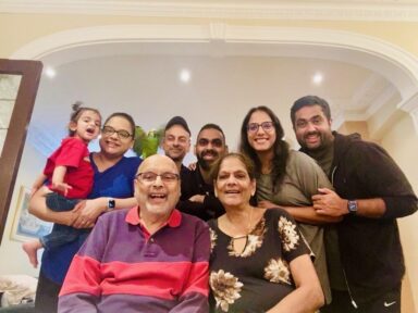 A family of eight, including a toddler, sitting and standing in a living room and smiling at the camera.