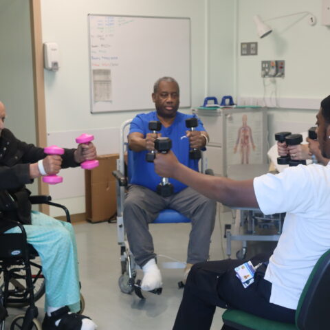 Two male patients sat in wheelchairs with weights in their hands. Two physiotherapists sat nearby also holding weights.