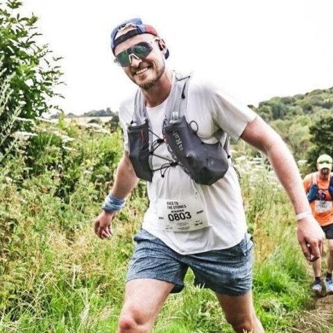 Man in full running gear and sunglasses smiling and running through the field with another runner behind him.
