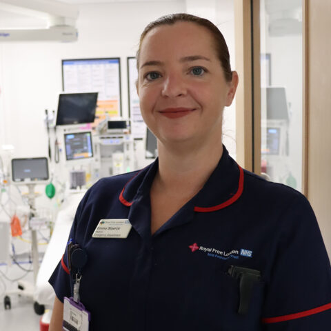 A female nurse wearing a navy blue Royal Free London NHS uniform stands smiling in a hospital ward with medical equipment in the background.