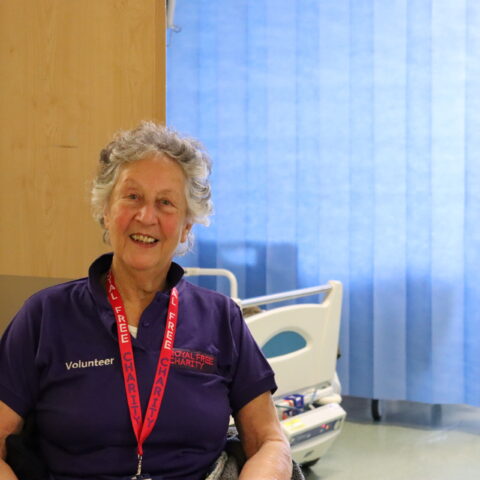A woman who is wearing a purple Royal Free Charity tshirt while sat in a wheelchair smiling.