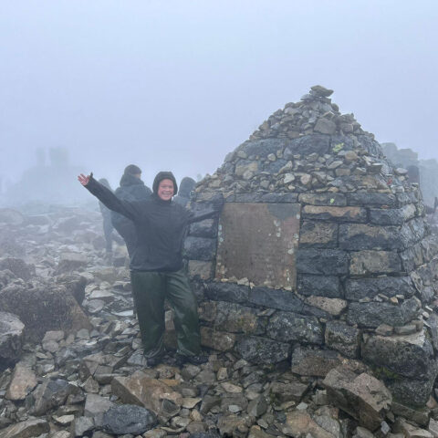 A woman in outdoor trekking grear standing beside a small hut made of bricks and rocks on the top of a misty mountain.