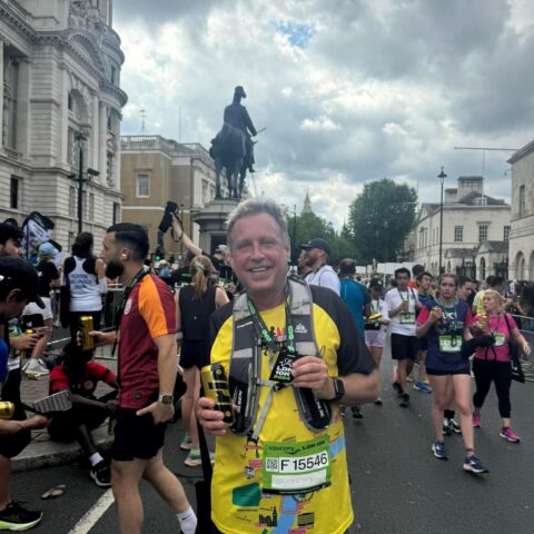 Man standing in the rood in running gear wearing a finishes medal, surrounded by other runners.