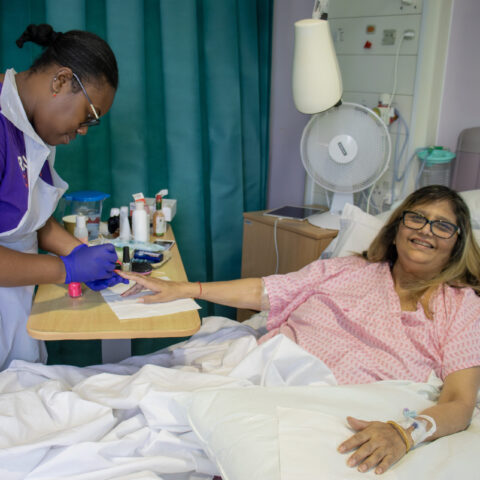 A female beautician, who is wearing a Royal Free Charity branded t-shirt, painting the nails of a woman who is lay in a hospital bed.