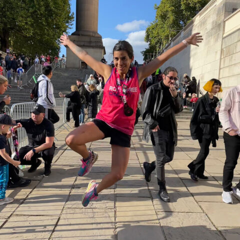 A woman in a Royal Free Charity vest vest jumping in the air with her hands raised.
