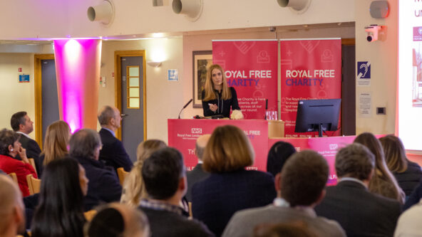 A woman standing in front of a lectern with Royal Free Charity branding, in front of an audience of 100 people.