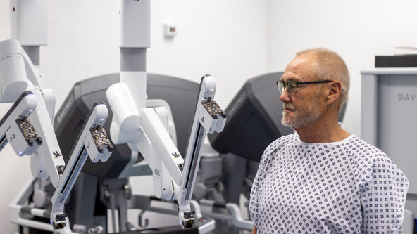 A patient in a hospital gown and glasses standing beside the new surgical robot.