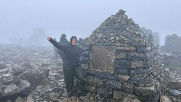 A woman in outdoor trekking grear standing beside a small hut made of bricks and rocks on the top of a misty mountain.