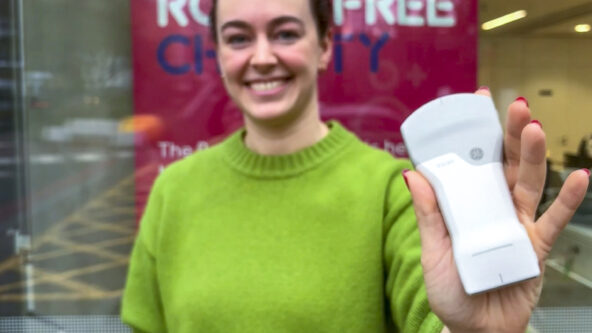 A woman in a green jumper is holding a pocket-sized ultrasound scanner towards the camera.