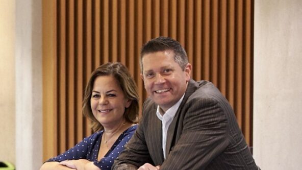 A smartly-dressed woman and man are leaning on a glass railing, smiling at the camera.