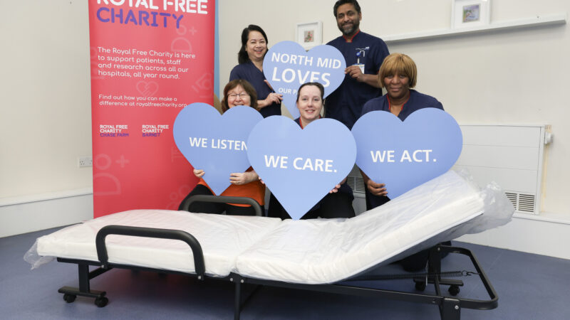 Five people holding large blue hearts - with words on them - standing and kneeling in front of a portable bed. Beside them all is a Royal Free Charity banner.