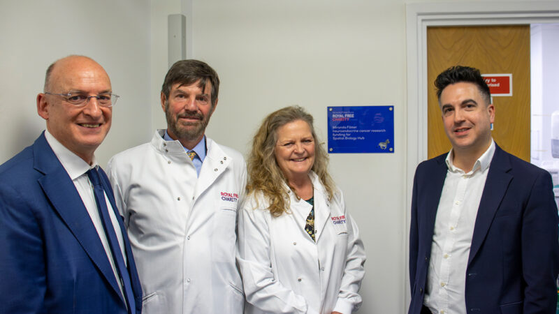 Four people standing in a row, two wearing Royal Free Charity lab coats, by a plaque on the wall that reads: Miranda Filmer neuroendocrine cancer research funding for Spatial Biology Hub.