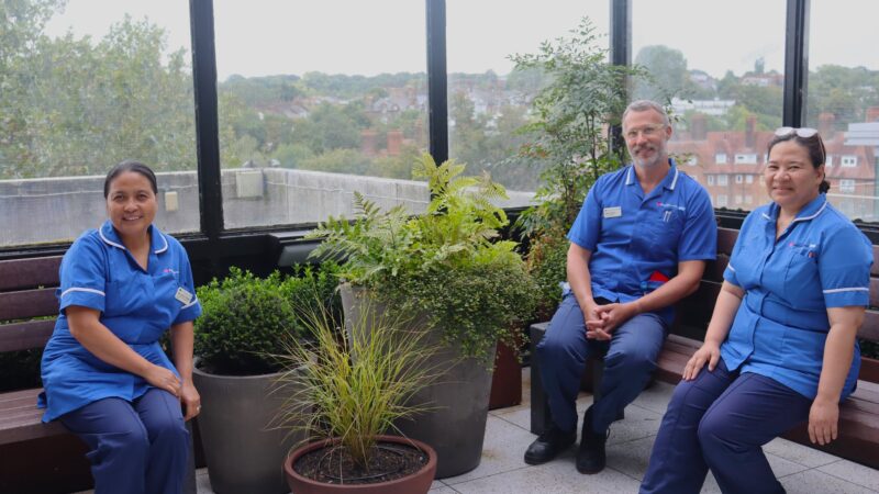 Three members of NHS wearing blue uniforms sat on benches in an outdoor garden.