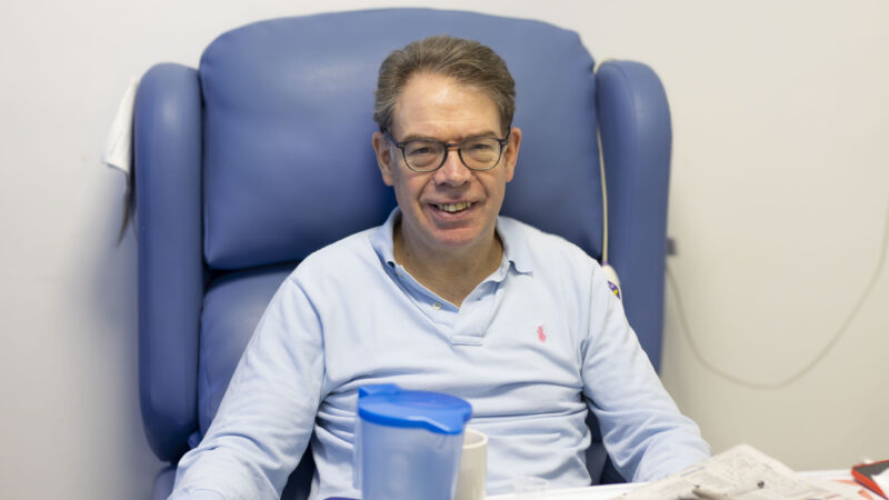 A man sat in a blue hospital chair with a desk in front of him. He is wearing a blue, long-sleeved t-shirt and glasses and is smiling.
