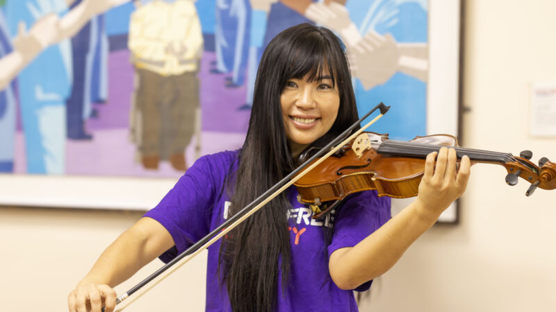 A woman with long dark hair wearing a Royal Free Charity t-shirt is playing the violin and smiling at the camera.