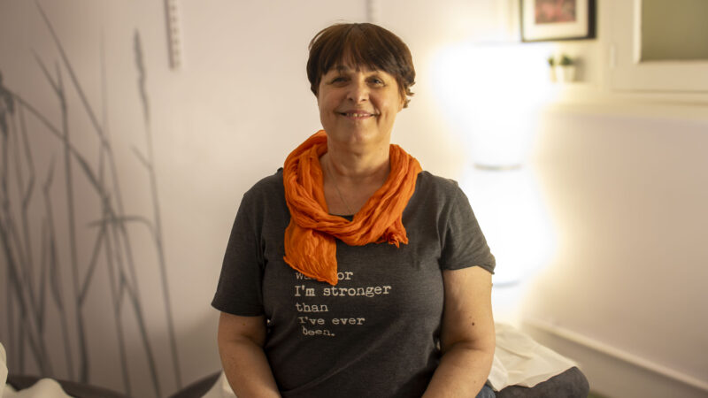 A woman with short brown hair smiles while sitting cross-legged on a bed or cushion in a softly lit room. She is wearing a dark grey t-shirt with text and an orange scarf around her neck.