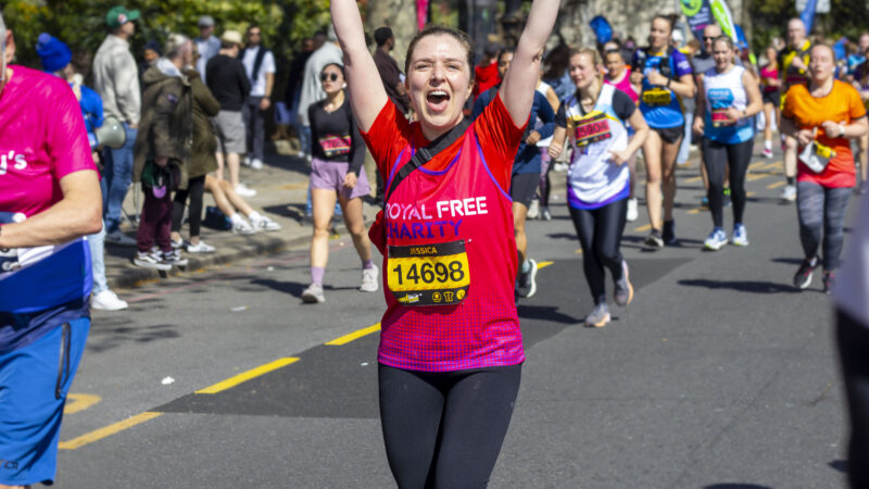 A runner in a Royal Free Charity top celebrates with arms raised while crossing the finish line at a road race, with other runners behind.