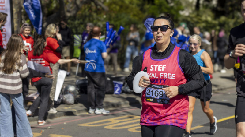 A runner wearing a pink Royal Free Charity vest takes part in a road race, holding a water bottle as spectators cheer from the sidelines.