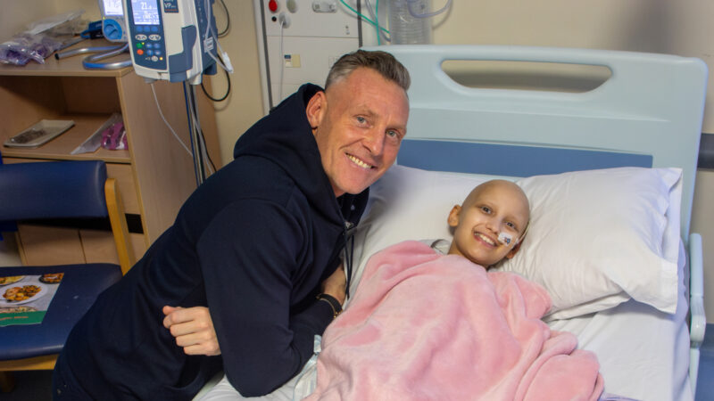 A man is crouched next to a young girl who is lying in a hospital bed wrapped in a pink blanket. Both are smiling.