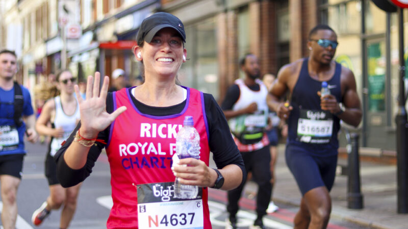 A runner wearing a Royal Free Charity vest smiles and waves while running in a city road race, holding a water bottle.