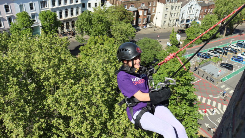 A zoomed out photo of a woman wearing a Royal Free Charity tshirt doing an abseil down the Pears Building in Hampstead.