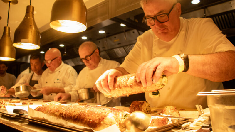 Three male chefs wearing glasses lined up at a restaurant pass. The chef closest to the camera is moving meat from one plate to another.