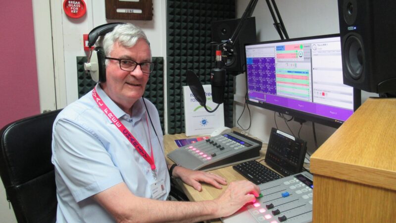A man sat at a radio desk with a computer and microphone in front of him.